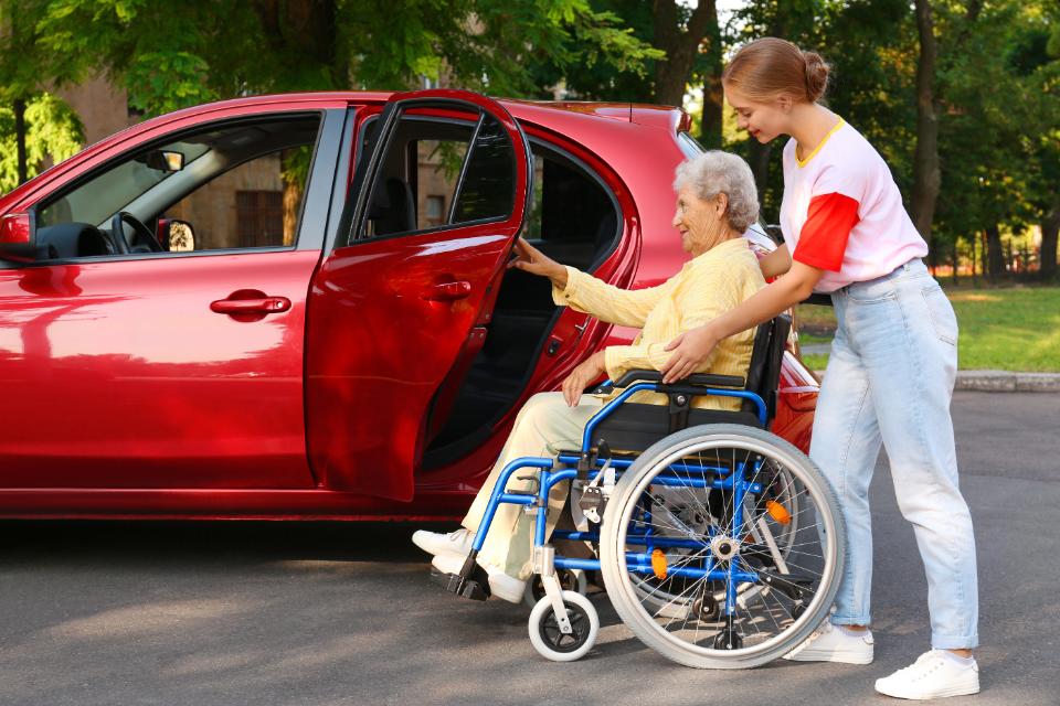 Caregiver supporting elderly lady on wheelchair to get into the car